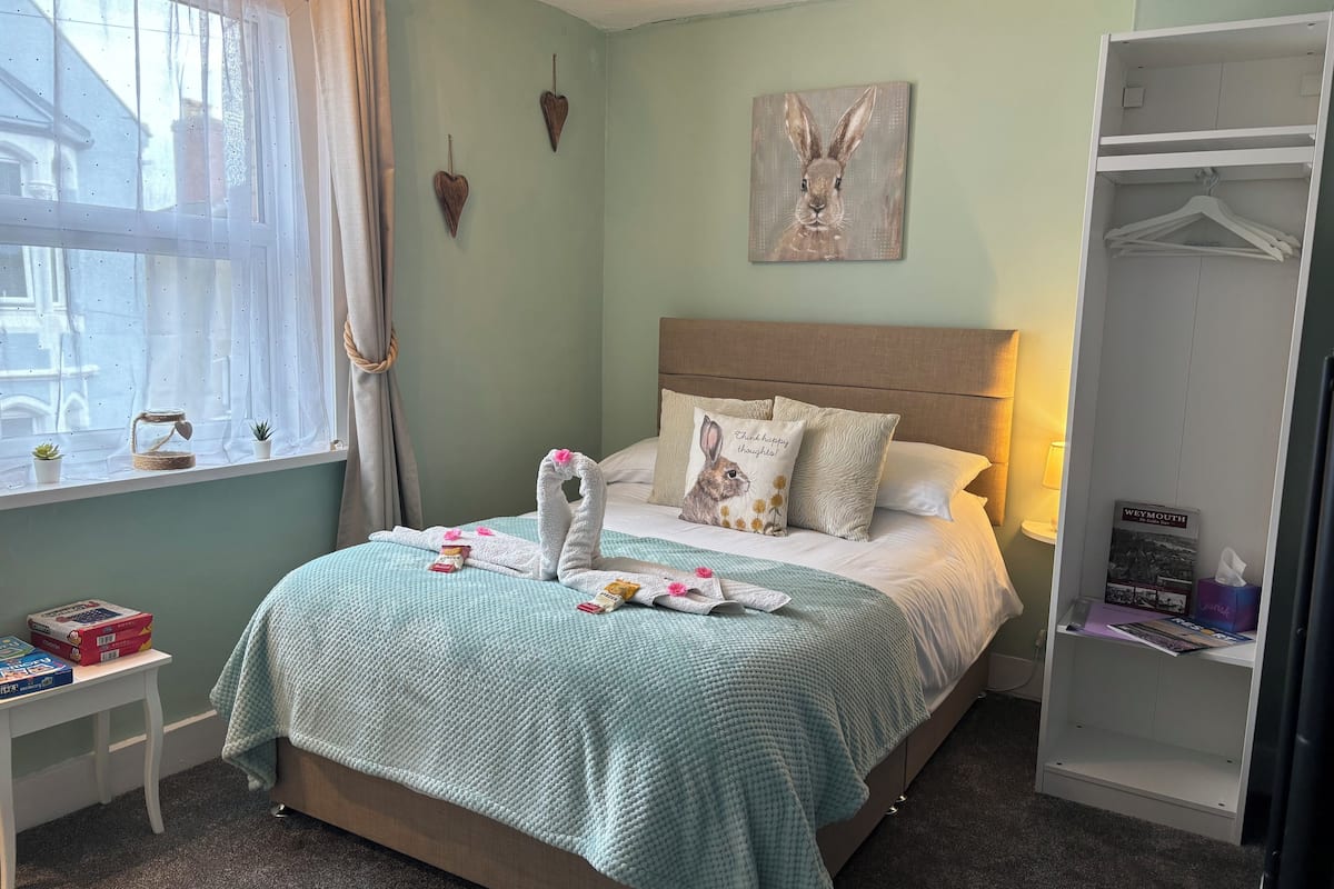 A neatly made bed with a teal blanket, a pillow with a rabbit design, and a heart-shaped towel arrangement. A small table with a plant and a candle holder is beside the bed.