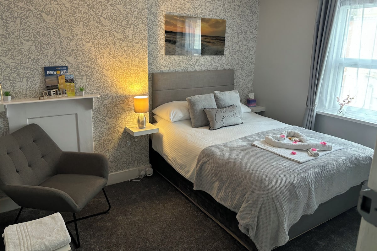 A hotel room with a bed, a chair, a nightstand with a lamp, a picture on the wall, and a window with curtains.