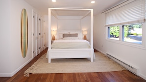 Een wit hemelbed met een beige vloerkleed, houten vloer en een raam met zonwering.