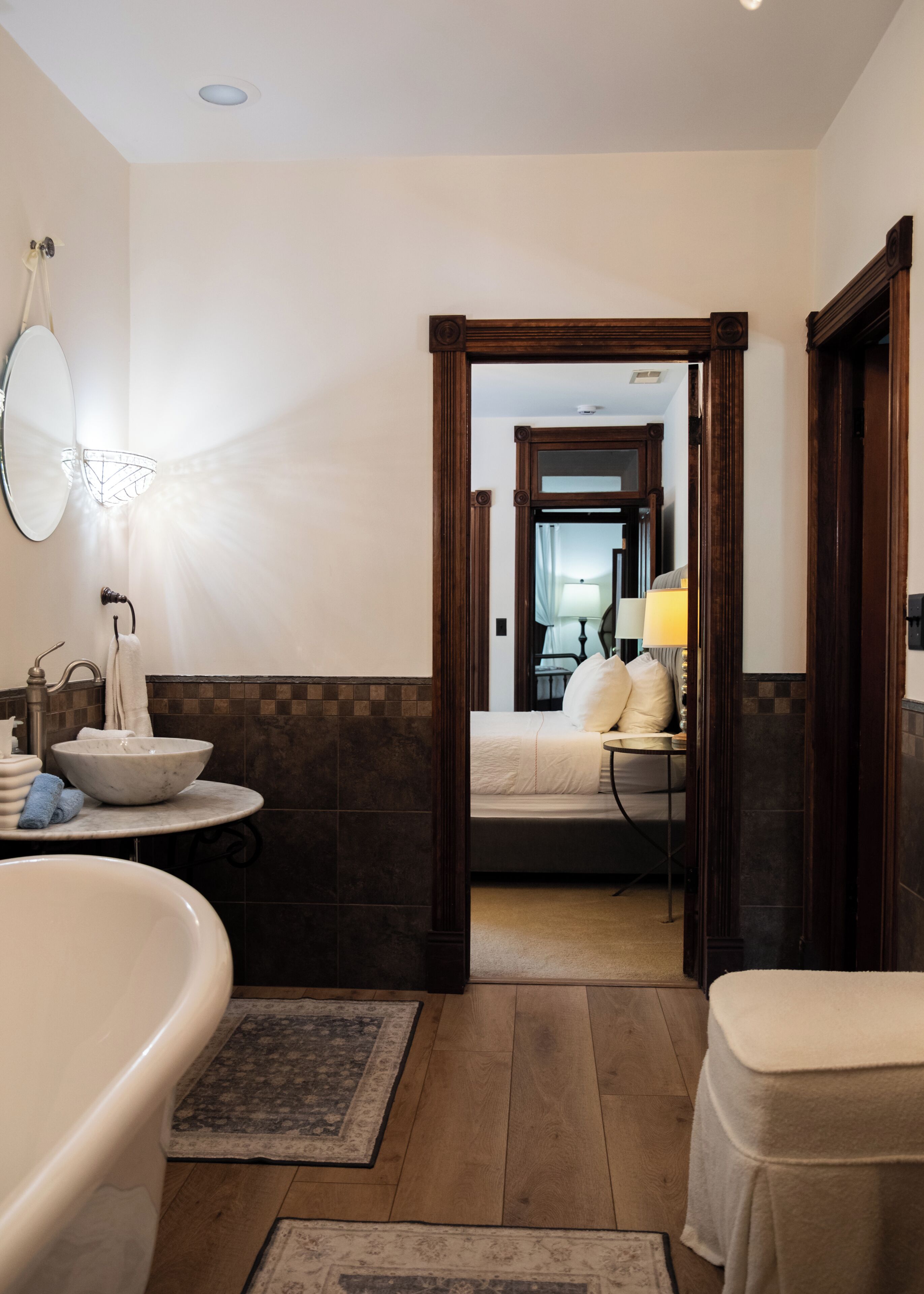 A bathroom with a bathtub, sink, and mirror. There is a doorway leading to a bedroom with a bed and a nightstand.