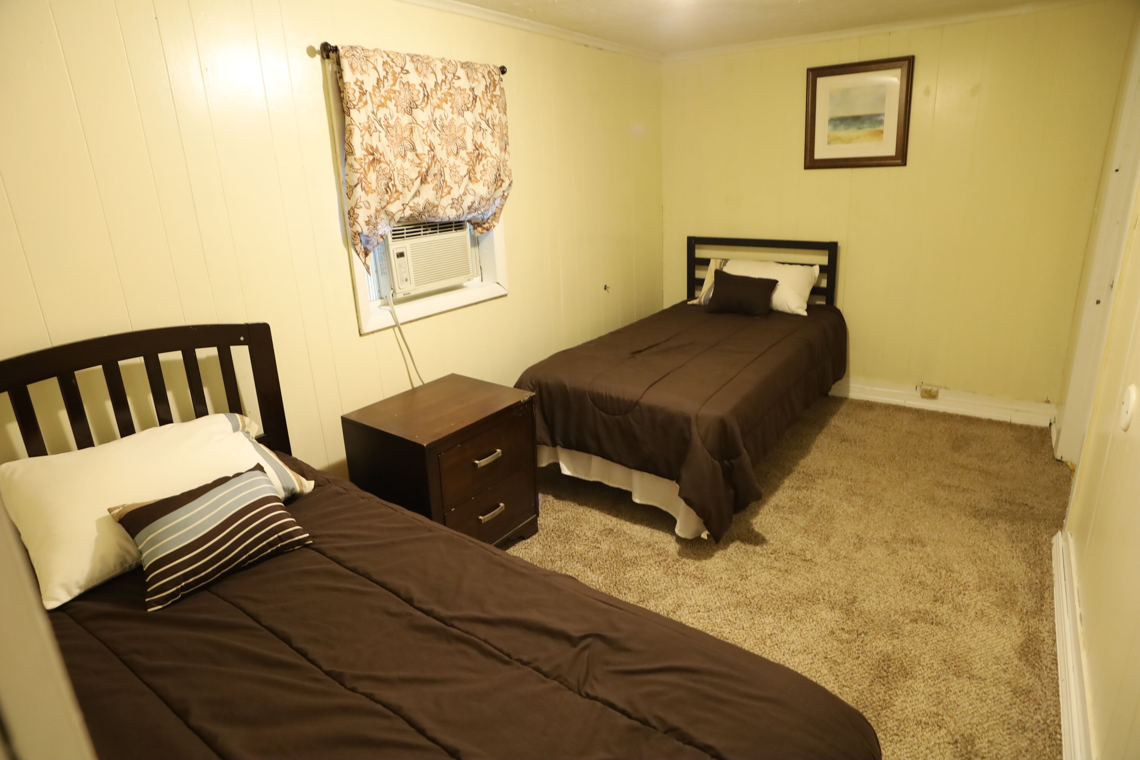 A bedroom with two single beds, a nightstand, a dresser, a window with a curtain, and a framed picture on the wall.