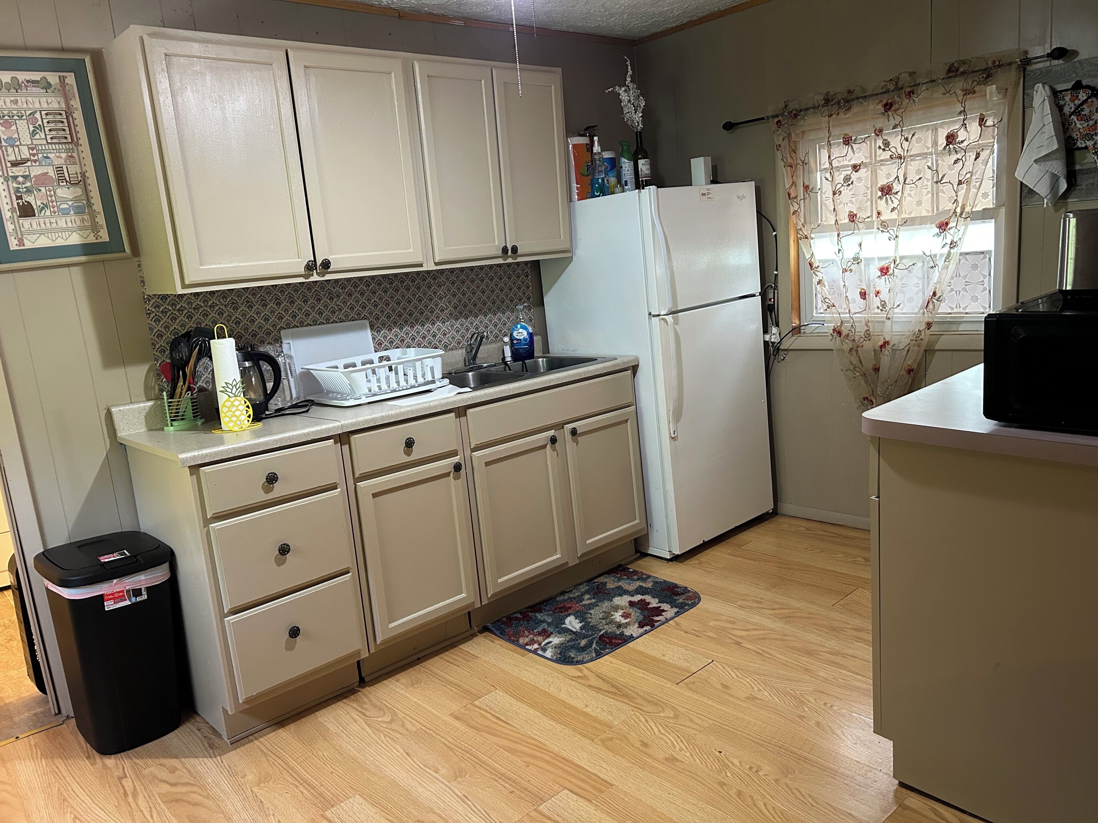 A kitchen with light-colored cabinets, a refrigerator, a sink, and a trash bin.