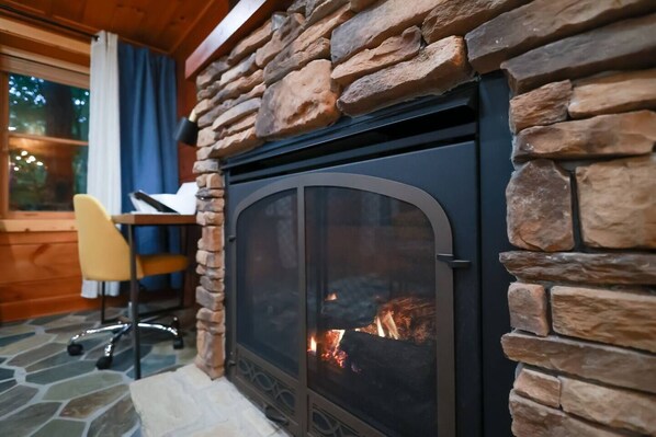 A stone fireplace with a glass door, a wooden desk, and a chair in a room with wooden walls.