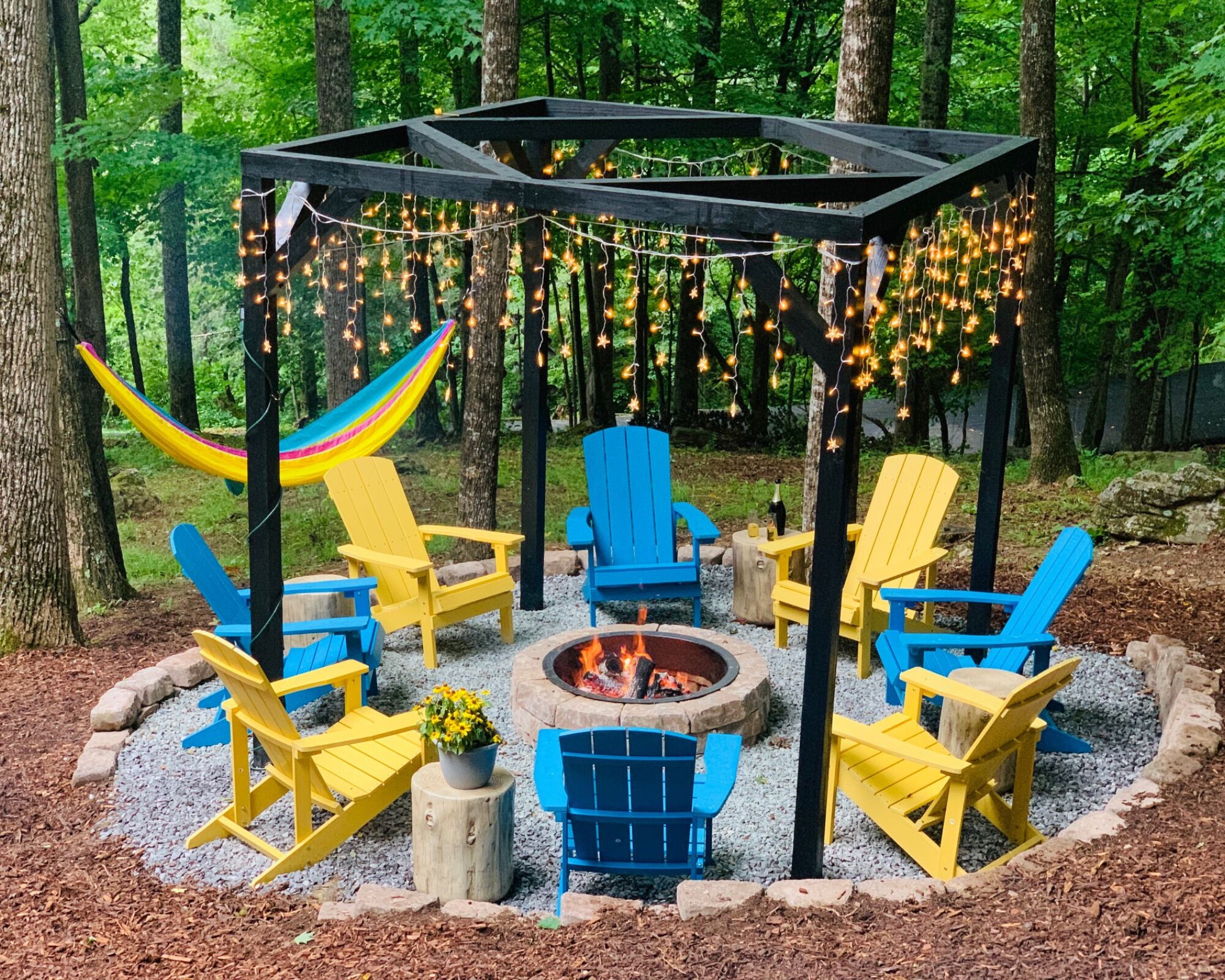A fire pit surrounded by yellow and blue Adirondack chairs, with a hammock and string lights in a forest setting.