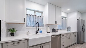 A modern kitchen with white cabinetry, a stainless steel refrigerator, a dishwasher, and a window with a curtain.
