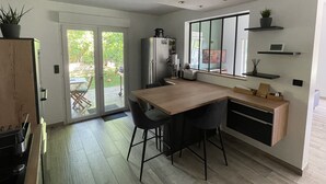 Une cuisine moderne avec une table en bois, deux chaises, un réfrigérateur et une fenêtre surmontée d’étagères.