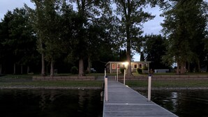 Eine Holzsteg, der zu einem Haus mit Veranda führt, umgeben von Bäumen und ruhigem Gewässer.