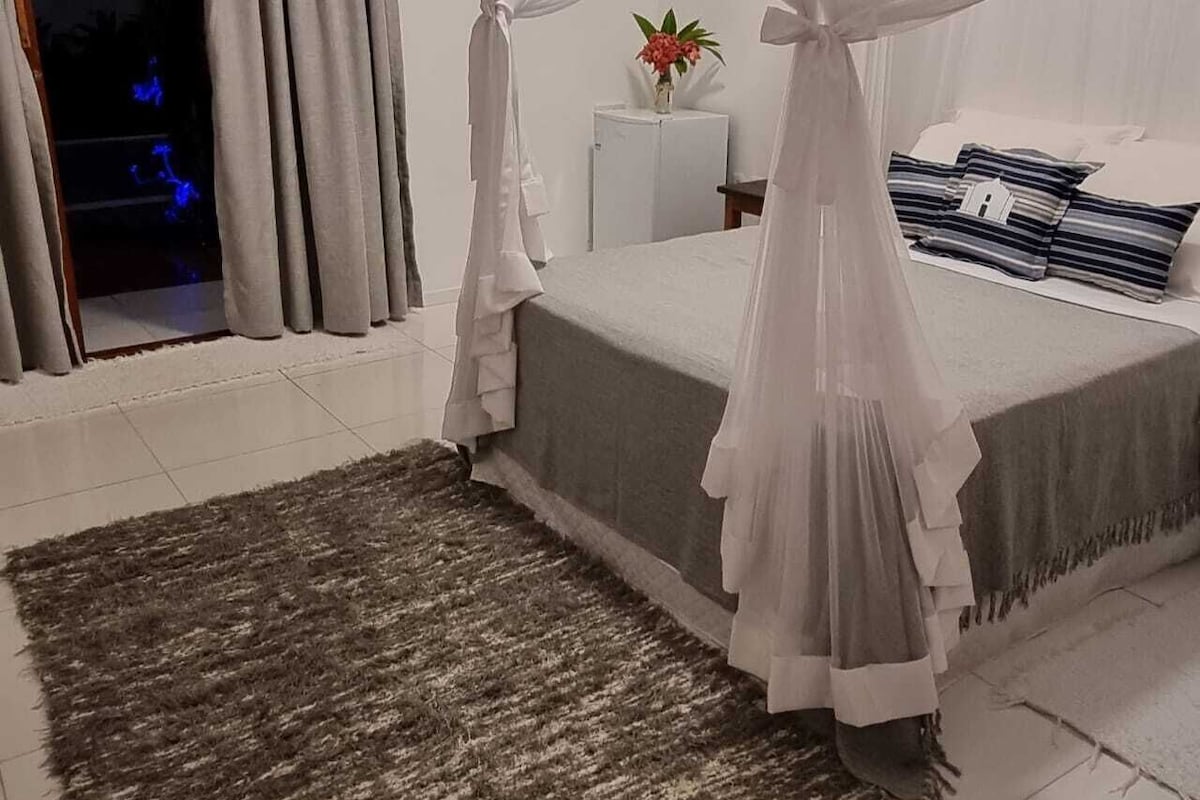 A bedroom with a bed, a rug, a nightstand, a plant, and a window with curtains.