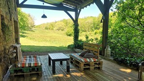 A wooden deck with a bench and a table, surrounded by greenery and a view of a grassy field.