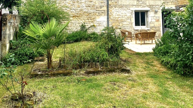 Une maison en pierre avec un petit jardin, un palmier et une terrasse meublée de mobilier en osier.