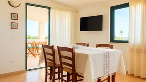 A dining area with a table set for four, wooden chairs, a white tablecloth, a flat-screen TV mounted on the wall, and a view of the outdoors through a window. - Panoramic Villa Sea View  (Quartu Sant'Elena)