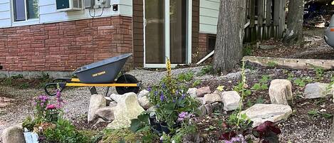 A garden with various potted plants and rocks, a wheelbarrow, and a house with a brick facade and a sliding glass door.