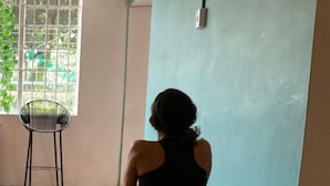Eine Person führt eine Yoga-Pose auf einer grünen Matte in einem Raum mit Holzboden und einem Fenster mit Blick auf Grünflächen.