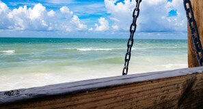 A wooden swing with metal chains, overlooking a beach with clear turquoise water and a cloudy sky.