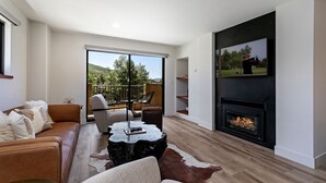 A modern living room with a fireplace, a large TV, and a view of a balcony with outdoor furniture. - Modern Condo in Avon, Walk to BC Gondola, SA228 (Avon)