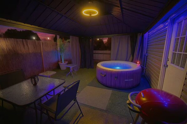 Ein Whirlpool in einem überdachten Außenbereich mit Tisch und Stühlen, umgeben von einer Holzwand und einem Fenster.