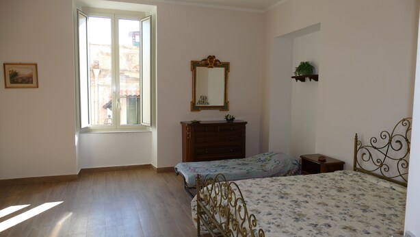 A bedroom with a wooden floor, a bed with a floral bedspread, a dresser, a mirror, and a window with a view of buildings outside.