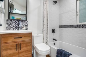 A bathroom with a white toilet, a wooden vanity with a mirror, a bathtub, and patterned tiles. - Ocean View Getaway! (Long Beach)