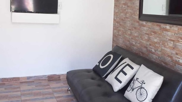 A modern living room with a black leather sofa, a mounted TV, a clock, and a mirror on the wall.