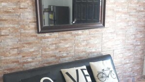A black leather sofa with two cushions, one with "LOVE" and another with a bicycle design, against a brick-patterned wall with a mirror above.