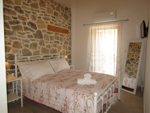 Un dormitorio con pared de piedra, cama con ropa de cama floral, mesita de noche, lámpara y espejo.