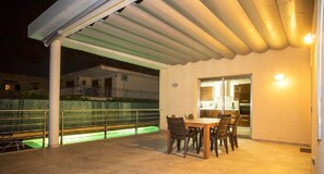 A covered outdoor patio with a dining table and chairs, a pool with green lighting, and a view of a residential area at night.