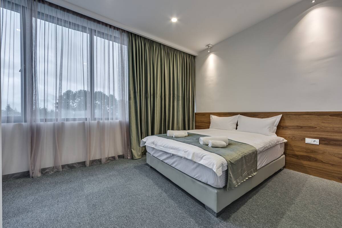 A modern hotel room with a large bed, grey carpet, wooden headboard, and sheer curtains.