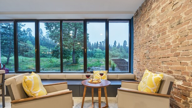 A cozy seating area with two chairs and a small table, a brick wall, and a large window overlooking a forested area. - Mountain Resort Gantiadi (Borjomi)