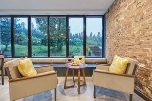 Un coin salon accueillant avec deux fauteuils et une petite table, un mur en briques et une grande fenêtre donnant sur une zone boisée.