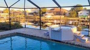 En swimmingpool med jacuzzi og liggestole, omgivet af et glasafskærmet område med udsigt til huse og et vandareal.