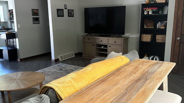 Un salon avec une table à manger en bois, des canapés gris, un téléviseur à écran plat et une bibliothèque.