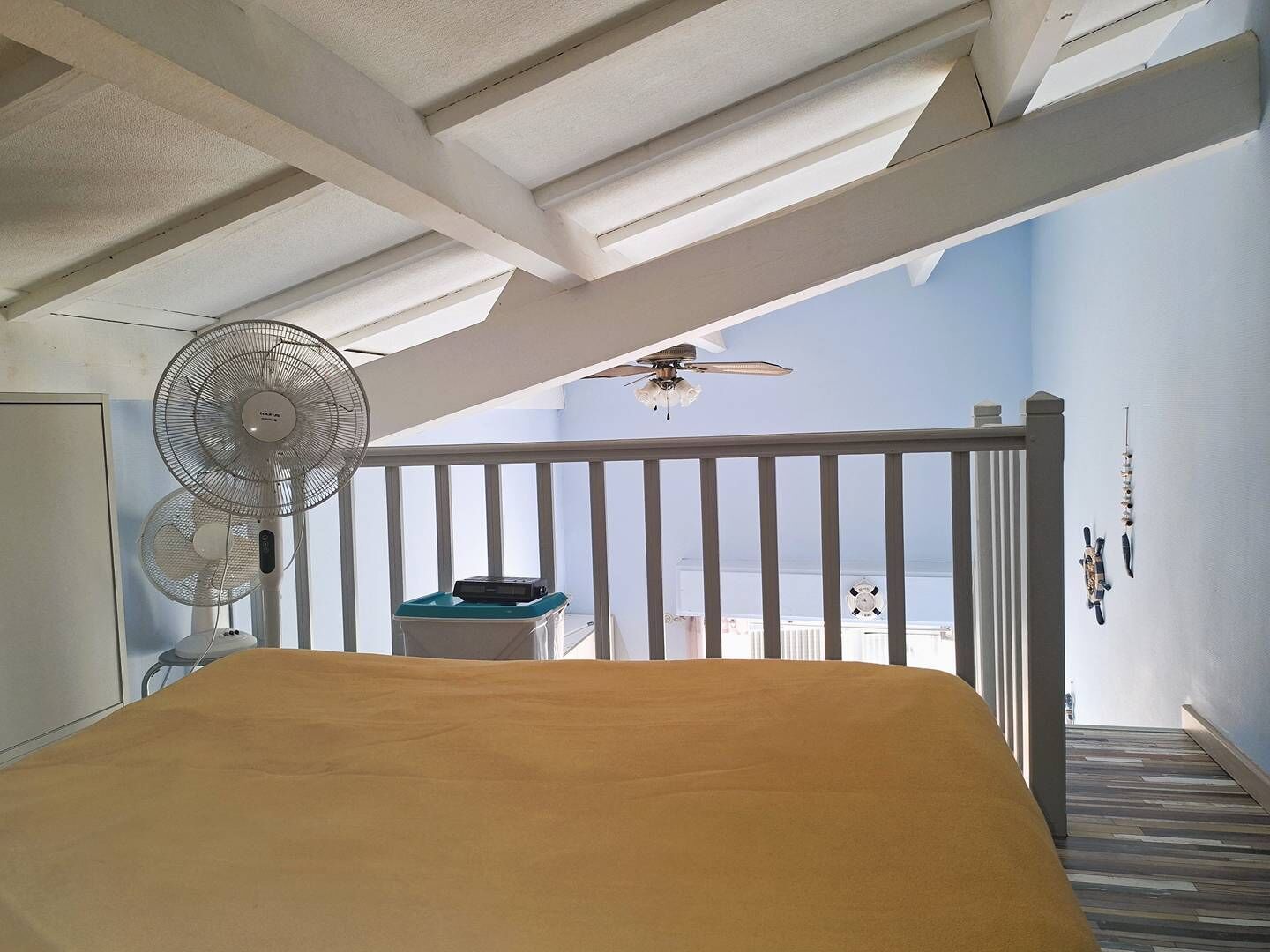 A bedroom with a bed, a ceiling fan, a small table with a radio, and a balcony with a view of the sea.