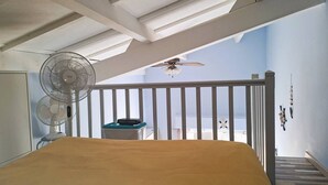 A bedroom with a bed, a ceiling fan, a small table with a radio, and a balcony with a view of the sea.