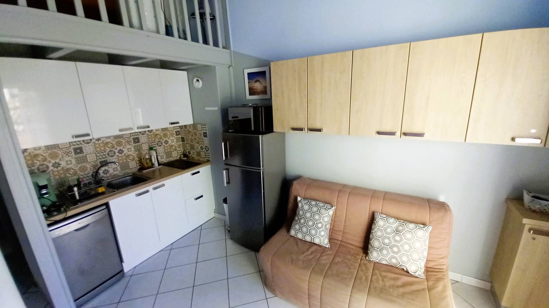 A compact kitchen with white cabinets, a refrigerator, and a microwave. There's a brown sofa with patterned cushions in the living area.