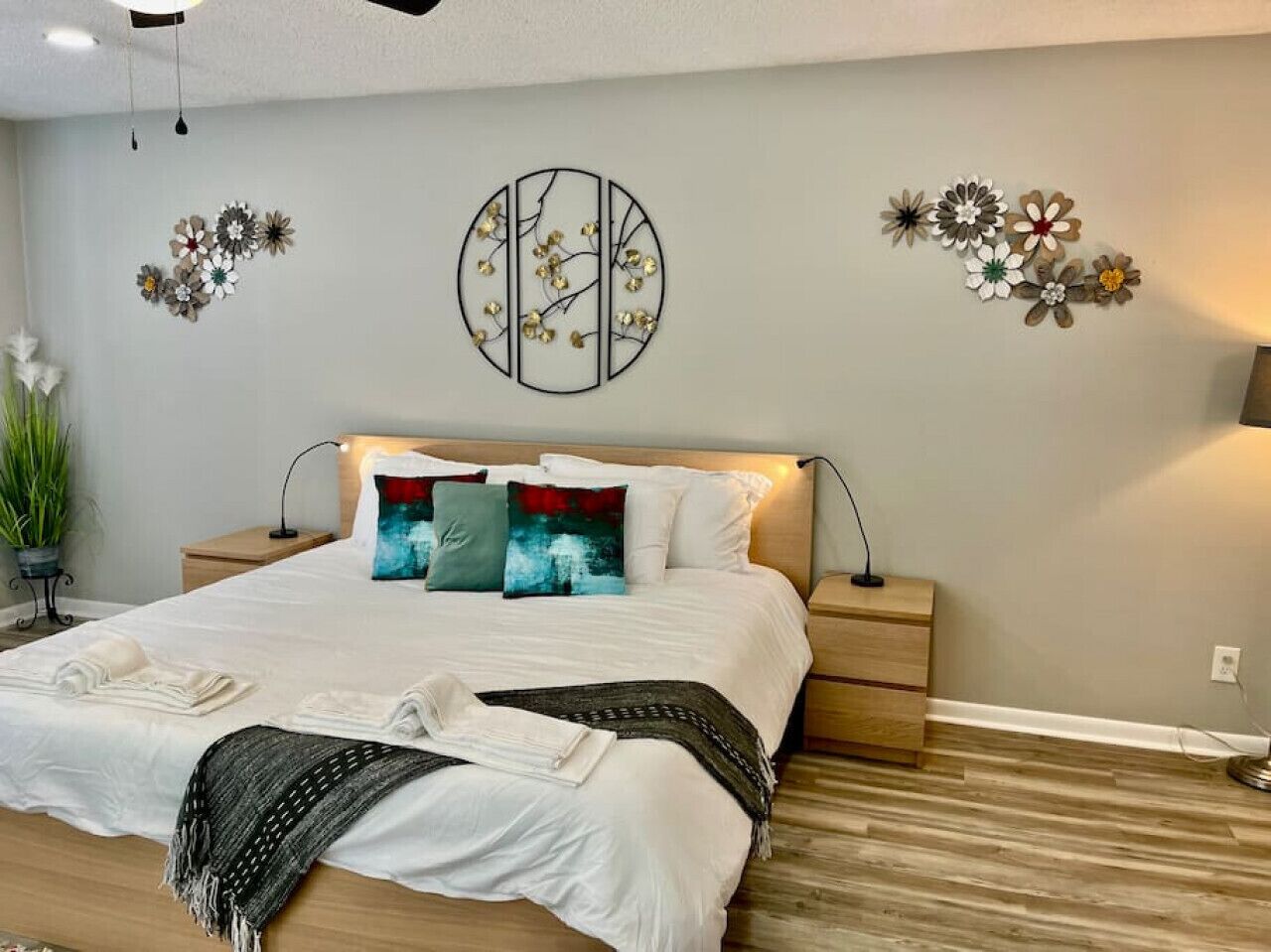 Una cama bien hecha con sábanas blancas y almohadas decorativas, una mesita de noche con lámpara y una obra de arte en la pared sobre la cama.