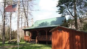 Een houten blokhut met een veranda, omringd door bomen en een vlag.