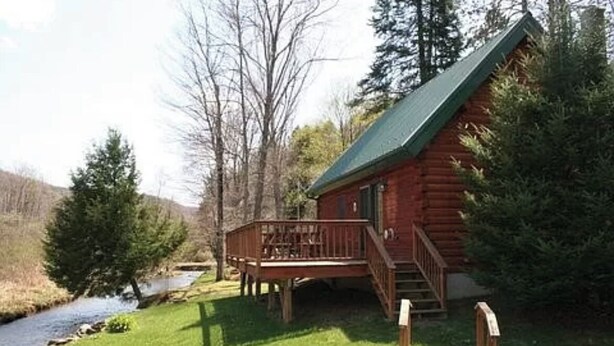 Een houten blokhut met een terras en een kleine veranda, omgeven door bomen en een rivier.
