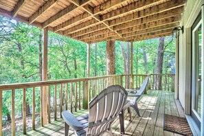 A covered outdoor deck with wooden railings, two Adirondack chairs, and a view of a forested area.