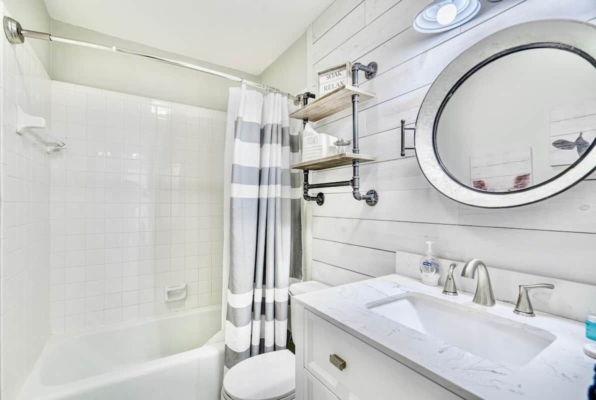 A modern bathroom with a white bathtub, a shower curtain, a toilet, a sink with a mirror, and a shelf with toiletries.