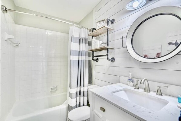 A modern bathroom with a white bathtub, a shower curtain, a toilet, a sink with a mirror, and a shelf with toiletries.