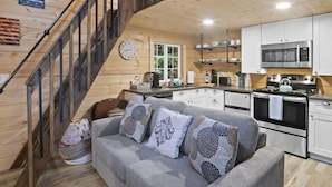 A cozy cabin interior with a staircase, grey sofa with patterned cushions, wooden walls, and a kitchen area with modern appliances. - Tiny Home A, just steps from Mt. Rainer Nat. Park w/firepit. (Ashford)