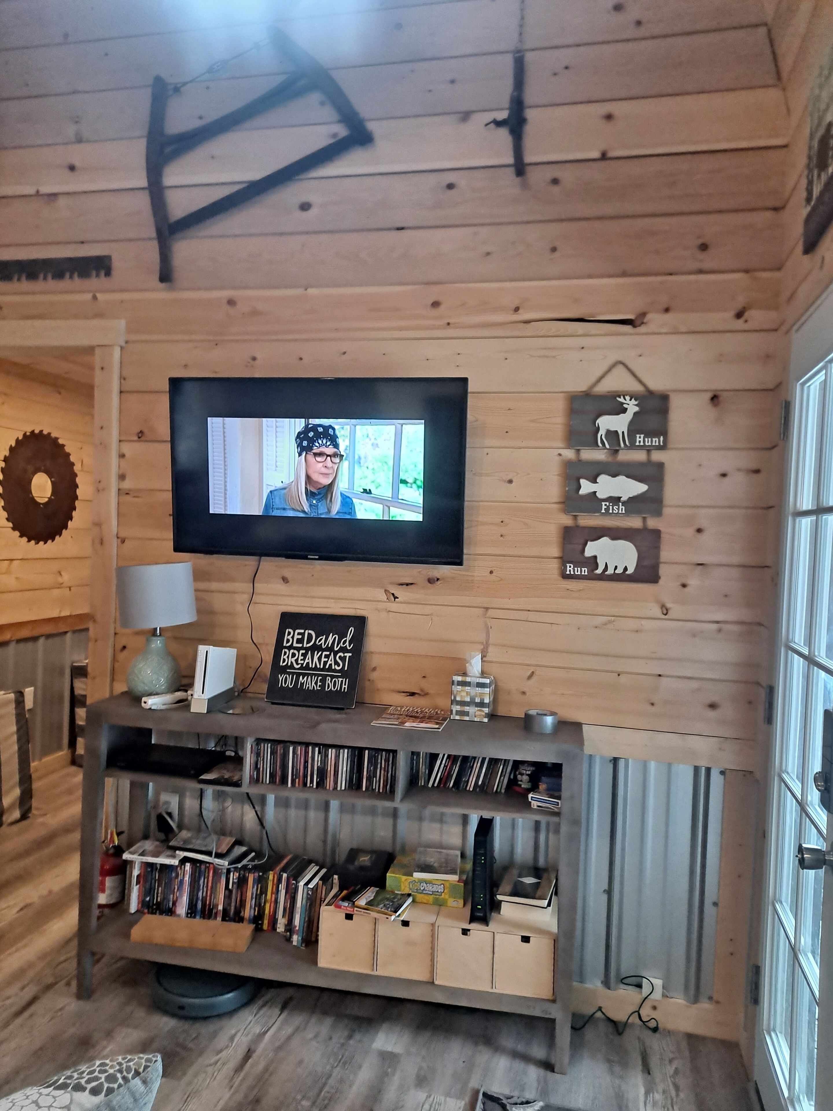 A cozy room with a television mounted on a wooden wall, a shelf with books and a sign saying "BEDand BREAKFAST YOU MAKE BOTH", and a window with a view of greenery.
