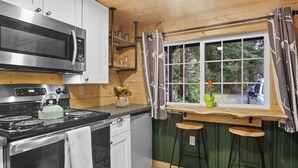 A kitchen with a microwave, stove, and sink. There are stools at a window with a view of trees. - Tiny Home A, just steps from Mt. Rainer Nat. Park w/firepit. (Ashford)