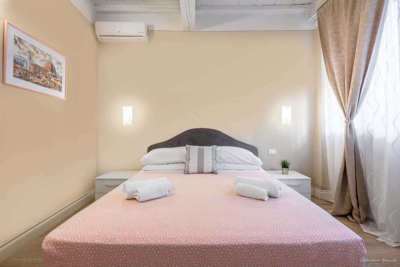 A neatly made bed with a pink patterned bedspread, two pillows, and two folded towels. There is a small bedside table with a plant, a framed picture on the wall, and a window with curtains.
