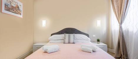 A neatly made bed with a pink patterned bedspread, two pillows, and two folded towels. There is a small bedside table with a plant, a framed picture on the wall, and a window with curtains.