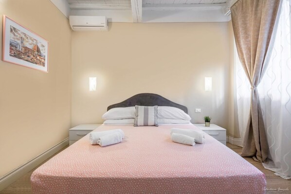 Una cama bien tendida con una colcha rosa estampada, dos almohadas y dos toallas dobladas. Se ve una mesita de noche con una planta, un cuadro enmarcado en la pared y una ventana con cortinas.