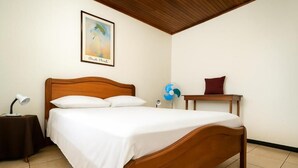 Een net opgemaakt bed met witte lakens, een houten hoofdbord, een klein nachtkastje met een lamp, een ventilator, en een rode kussens op een houten nachtkastje.