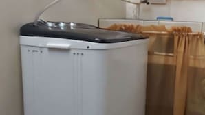 Een witte wasmachine in een hoek, met een betegelde vloer en een gordijn op de achtergrond.