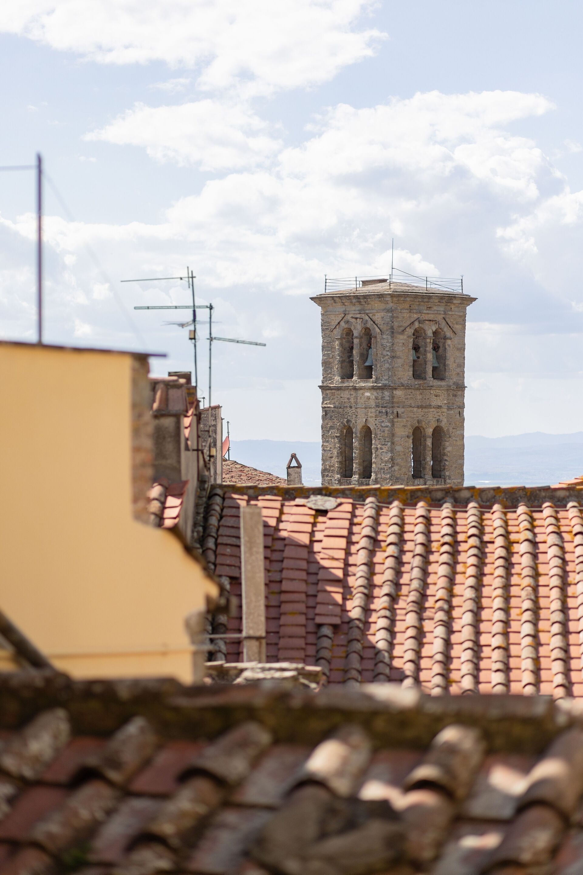 A historic tower with arched windows and a bell structure, viewed from a rooftop with terracotta tiles.