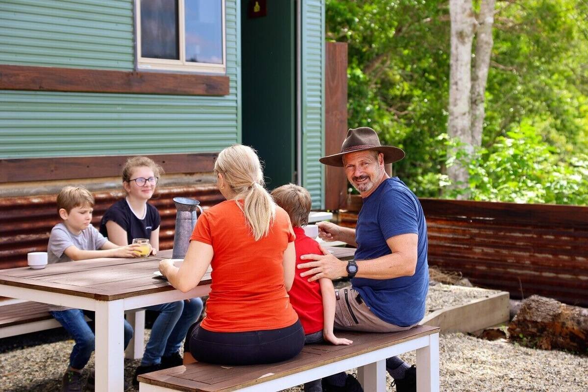 Una familia comiendo en una mesa de madera al aire libre, con un edificio verde al fondo.